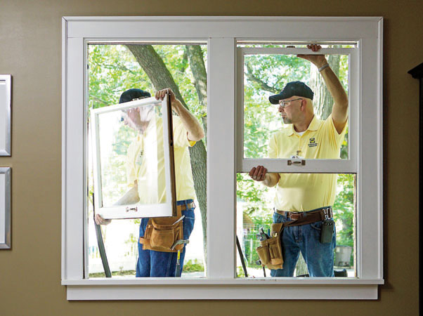 Two workers in yellow shirts installing a double-pane window in a residential home, with one person holding the window frame and the other securing the window in place. They are both wearing tool belts and are working together in a well-lit room with a view of trees outside.