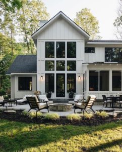 A modern two-story home with a pitched roof and large black-framed windows. The home features a high-quality roofing system with durable materials, perfectly complementing its white exterior. The spacious outdoor patio includes comfortable seating around a central fire pit, showcasing the home's seamless blend of stylish roofing and contemporary design amidst a lush green landscape.