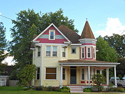 A charming Victorian-style house in Townsend, DE, featuring a light yellow exterior with red and white trim. The house has a prominent turret with a conical roof, a wrap-around porch, and large windows, creating a picturesque and welcoming appearance. Lush greenery surrounds the home, adding to its quaint and inviting atmosphere.
