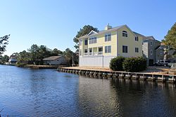 A picturesque view of a waterfront home in South Bethany, Delaware, featuring a light yellow house with a spacious balcony overlooking a calm canal on a clear day