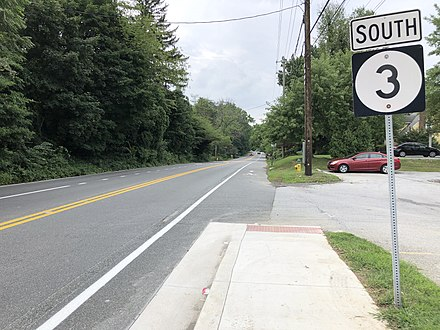 A road in Arden, Delaware, marked by a "South 3" road sign. The road is lined with dense trees on one side and a sidewalk on the other. A red car is parked near the sidewalk, and utility poles with overhead wires run alongside the road. The sky is overcast, adding a calm, quiet atmosphere to the scene.